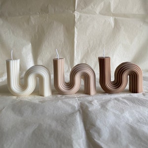 May include: Three wave-shaped candles in beige and brown hues. Each candle has a wick. The candles are arranged on a textured, off-white surface. The candles are a decorative item.