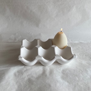 Könnte beinhalten: Ein weißer Keramik-Eierkarton mit einer einzelnen weißen Kerze in einem der Fächer. Die Kerze ist angezündet.