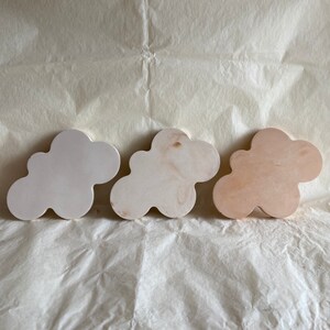 Peut inclure: Trois ornements en bois en forme de nuage dans différentes nuances de blanc et de beige. Les nuages sont disposés en rangée de gauche à droite, le nuage le plus clair à gauche et le nuage le plus foncé à droite.