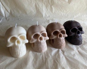 Può includere: Quattro candele a forma di teschio in diverse tonalità di marrone, bianco e nero. Le candele sono disposte in fila su una superficie marrone chiaro.
