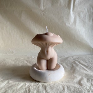 K&ouml;nnte beinhalten: Eine nude-farbene Kerze in Form eines menschlichen Torsos mit einem pilzf&ouml;rmigen Oberteil. Die Kerze steht auf einem wei&szlig;en runden Sockel.