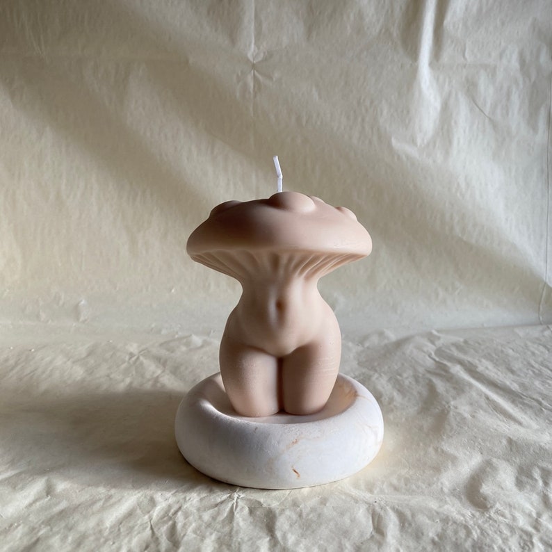 K&ouml;nnte beinhalten: Eine nude-farbene Kerze in Form eines menschlichen Torsos mit einem pilzf&ouml;rmigen Oberteil. Die Kerze steht auf einem wei&szlig;en runden Sockel.