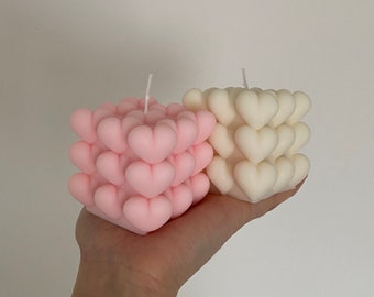 May include: Two heart-shaped candles, one pink and one white, are arranged in a cube shape. The candles are made of wax and have a smooth, glossy finish.