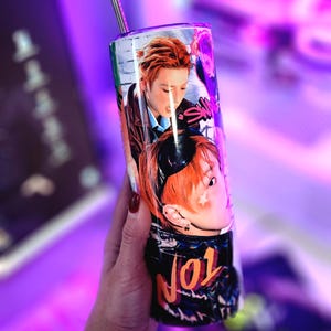 May include: A stainless steel tumbler with a straw, featuring a vibrant image of a person with orange hair. The tumbler has a black and orange design with the number "101" and other text. The background is a blurred purple.