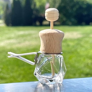 May include: A clear glass car air freshener with a geometric design. The bottle has a wooden top with a wooden stick inside. The bottle is attached to a white clip.