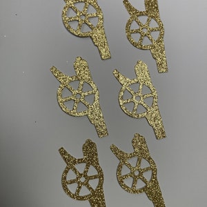 May include: Six gold glitter paper cutouts of a cannon with a wheel. The cannon is facing left and the wheel is in the center of the cutout.