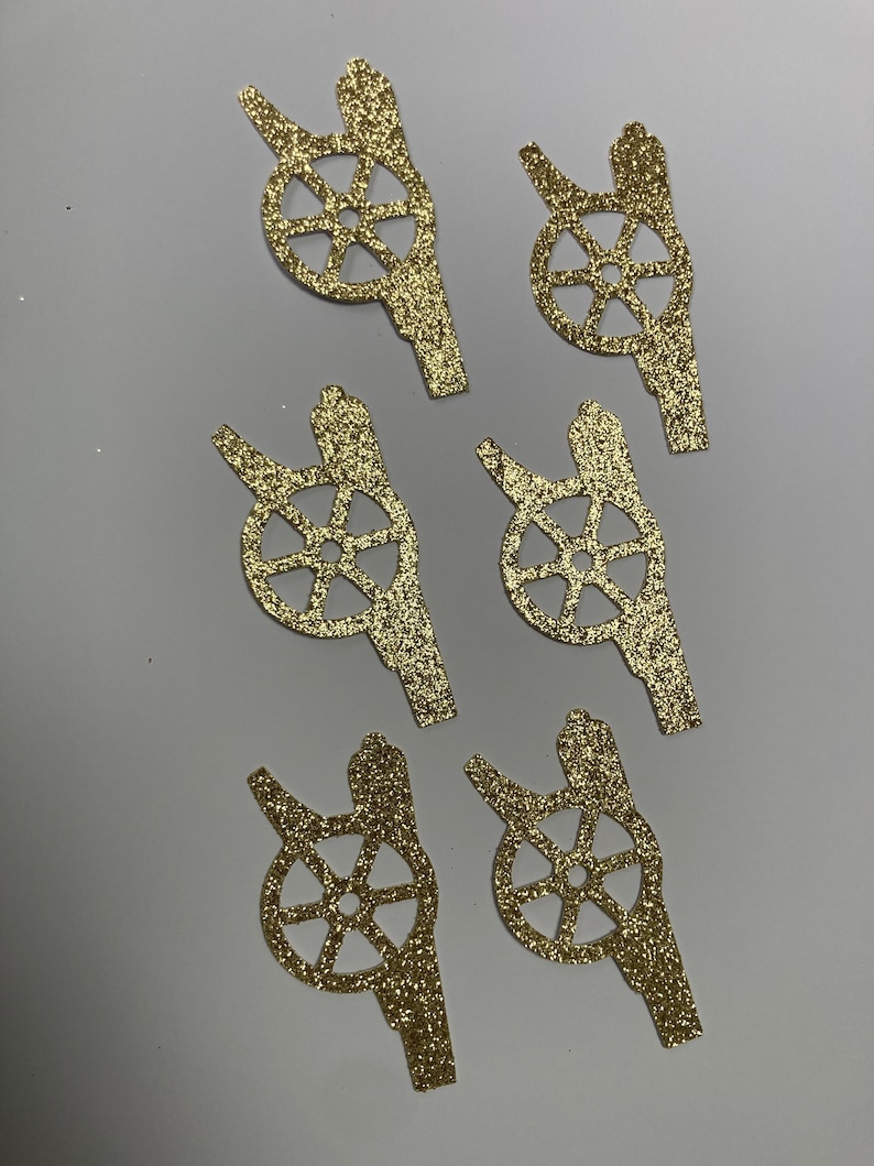 May include: Six gold glitter paper cutouts of a cannon with a wheel. The cannon is facing left and the wheel is in the center of the cutout.
