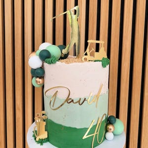 Puede incluir: Una tarta de cumplea&ntilde;os de dos pisos con tem&aacute;tica de golf. La tarta presenta glaseado verde y blanco, detalles dorados y un adorno para tarta de carrito de golf y golfista. El nombre "David" y el n&uacute;mero "40" tambi&eacute;n son dorados. Esferas decorativas verdes, blancas y doradas adornan la tarta.