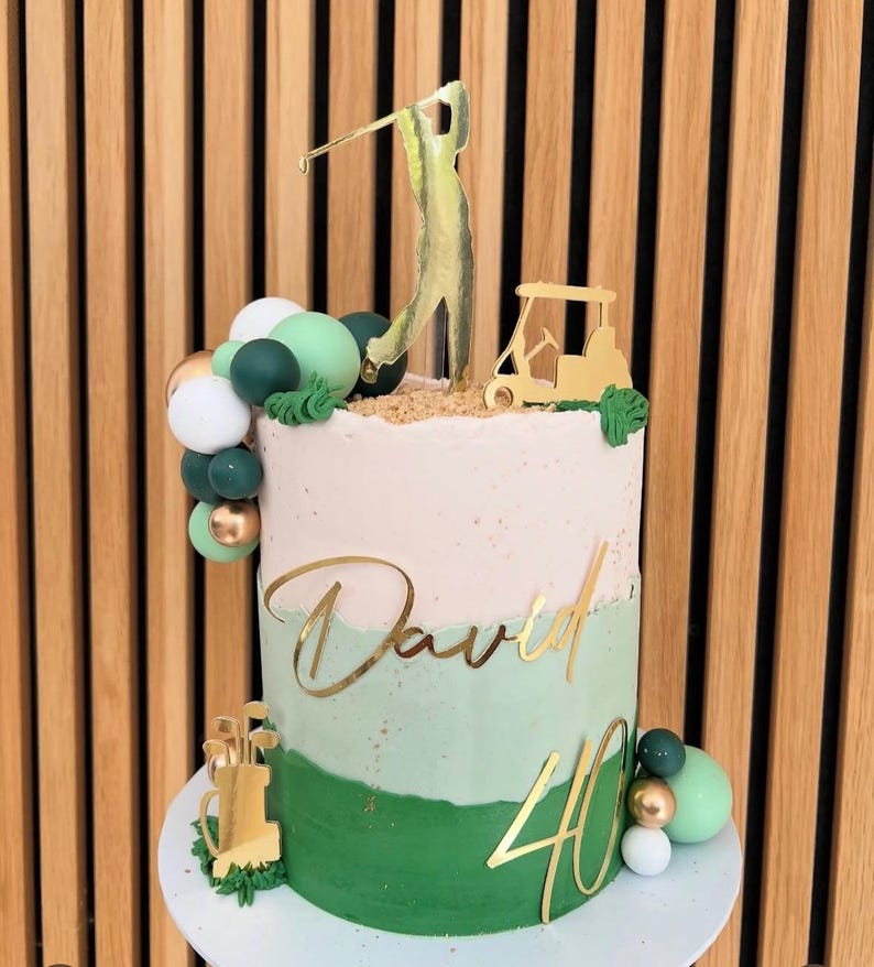 Puede incluir: Una tarta de cumplea&ntilde;os de dos pisos con tem&aacute;tica de golf. La tarta presenta glaseado verde y blanco, detalles dorados y un adorno para tarta de carrito de golf y golfista. El nombre "David" y el n&uacute;mero "40" tambi&eacute;n son dorados. Esferas decorativas verdes, blancas y doradas adornan la tarta.