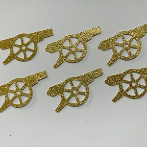 May include: Six gold glitter paper cutouts of a cannon with a wheel. The cannon is a common symbol for the Arsenal Football Club.