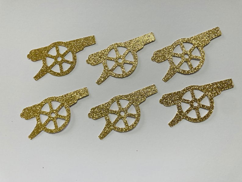 May include: Six gold glitter paper cutouts of a cannon with a wheel. The cannon is a common symbol for the Arsenal Football Club.