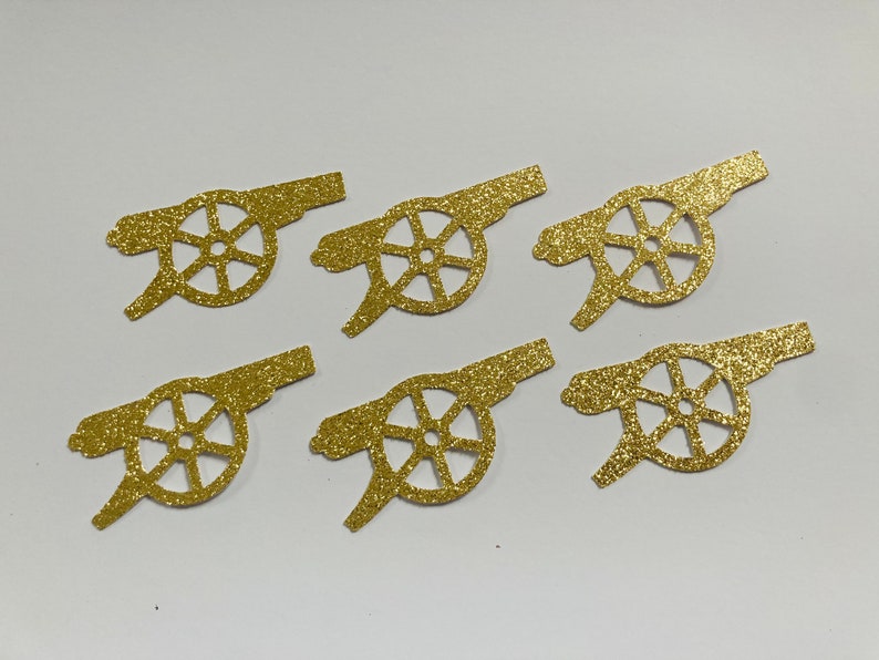 May include: Six gold glitter paper cutouts of a cannon with a wheel. The cannon is a common symbol for the Arsenal Football Club.