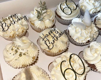 May include: A box of twelve cupcakes with white frosting. Some are decorated with "Happy Birthday" or "60" toppers, while others have sprigs or gold sprinkles. A selection of celebratory cupcakes, perfect for a birthday celebration.