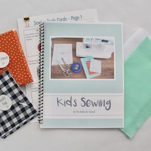 Op de afbeelding: Een naaiset voor kinderen, met een spiraalgebonden boek getiteld "Kid's Sewing". Inclusief oranje stof met witte stippen, een zwart-wit geruite voorraadzak en vilt.