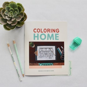 May include: A coloring book titled "Coloring Home" with a black and white illustration of a house on the cover. The book is on a white surface with two pencils, a pencil sharpener, and a small succulent plant.