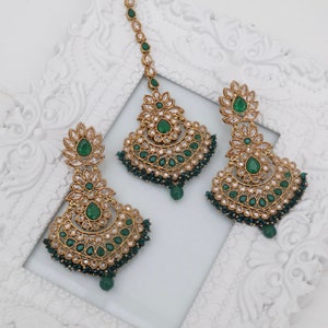 May include: A set of gold-tone earrings and a matching maang tikka with green and clear stones. The jewelry is ornate and features a crescent moon design.