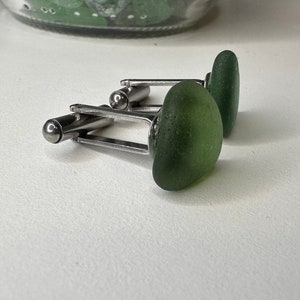 May include: A pair of cufflinks with green glass stones. The cufflinks are made of silver metal.