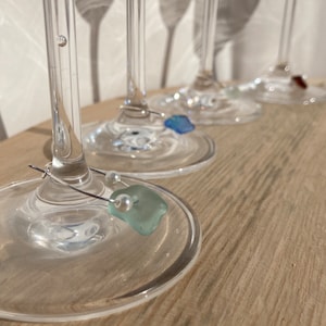 May include: Clear stemmed wine glasses with decorative charms. Each charm features a different coloured accent, including blue, green, and red sea glass, and a pearl. The glasses are arranged on a light wood surface.