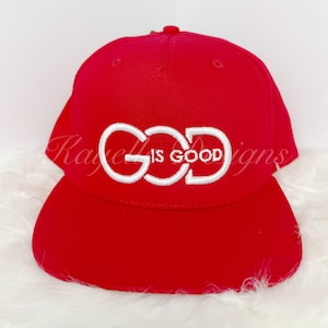 Op de afbeelding: Rode baseballpet met een wit geborduurd ontwerp met de tekst "GOD IS GOOD".