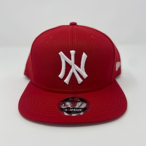 May include: A red and white New York Yankees baseball cap with a white embroidered logo and a black "New Era" logo on the front. The cap has a black adjustable strap with a white "Snapback" label.