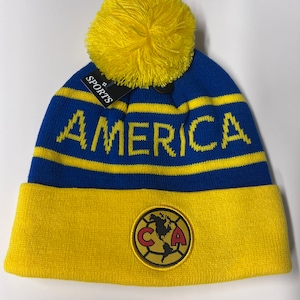 May include: Blue and yellow knit beanie with a yellow pom pom on top. The beanie has a yellow stripe and the word "AMERICA" in yellow letters. The beanie also has a yellow patch with a red "C" and "A" and a globe in the center.