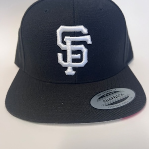 May include: Black baseball cap with a white embroidered "SF" logo. The cap has a black adjustable strap with a white "The Authentic Yupoong Snapback Yupoong Since 1974" label.