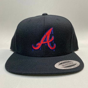 K&ouml;nnte beinhalten: Schwarze Baseballkappe mit einem roten und blauen "A"-Logo. Das Logo ist im Stil eines Tropfeneffekts gestaltet. Die Kappe hat einen schwarzen verstellbaren Riemen mit einem "The Authentic Yupoong Snapback"-Etikett.