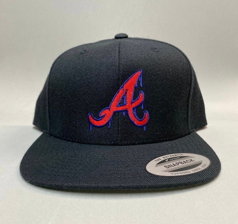K&ouml;nnte beinhalten: Schwarze Baseballkappe mit einem roten und blauen "A"-Logo. Das Logo ist im Stil eines Tropfeneffekts gestaltet. Die Kappe hat einen schwarzen verstellbaren Riemen mit einem "The Authentic Yupoong Snapback"-Etikett.