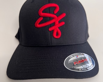 Könnte beinhalten: Eine schwarze Baseballkappe mit einem roten, gestickten Schriftzug auf der Vorderseite. Ein runder Aufkleber auf dem Schirm zeigt den Text THE ORIGINAL FLEXFIT L/XL IT FITS YOUR LIFE in weißer, roter und blauer Schrift auf silbernem Grund.