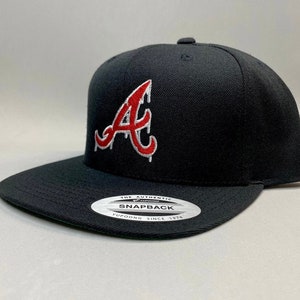 K&ouml;nnte beinhalten: Schwarze Baseballkappe mit einem roten und wei&szlig;en "A"-Logo. Die Kappe hat einen schwarzen Schirm und eine schwarze Krone. Das Logo ist ein stilisiertes "A" mit einem Tropfeffekt. Die Kappe hat ein wei&szlig;es Etikett auf der Unterseite, auf dem "The Authentic Yupoong Snapback Since 1974" steht.