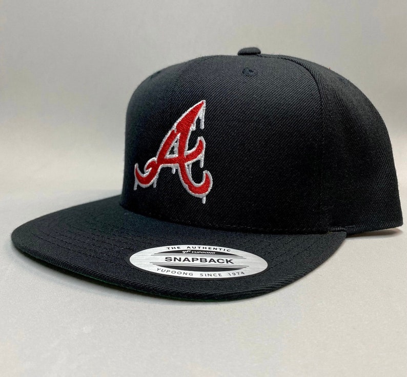 K&ouml;nnte beinhalten: Schwarze Baseballkappe mit einem roten und wei&szlig;en "A"-Logo. Die Kappe hat einen schwarzen Schirm und eine schwarze Krone. Das Logo ist ein stilisiertes "A" mit einem Tropfeffekt. Die Kappe hat ein wei&szlig;es Etikett auf der Unterseite, auf dem "The Authentic Yupoong Snapback Since 1974" steht.