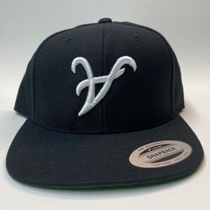 May include: A black and white baseball cap with a white embroidered letter 'V' on the front. The cap has a black brim and a black adjustable strap with a white 'Yupoong' logo and the text 'The Authentic Snapback Yupoong Since 1974'.