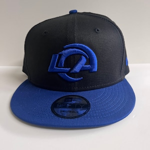 May include: A black and blue baseball cap with a blue "LA" logo on the front. The cap is a New Era 9Fifty Snapback.