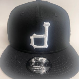 May include: Black and white baseball cap with a white "D" embroidered on the front. The cap is a New Era 9Fifty Snapback.