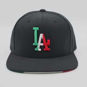 May include: Black baseball cap with a white, green, and red "LA" logo. The brim of the cap has a green, white, and red trim.