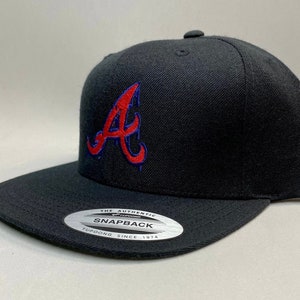 K&ouml;nnte beinhalten: Schwarze Baseballkappe mit einem roten und blauen gestickten "A" auf der Vorderseite. Die Kappe hat einen schwarzen Schirm und einen schwarzen Riemen mit einem wei&szlig;en Etikett "The Authentic Yupoong Snapback Since 1974".
