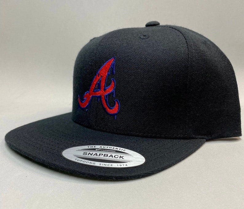 K&ouml;nnte beinhalten: Schwarze Baseballkappe mit einem roten und blauen gestickten "A" auf der Vorderseite. Die Kappe hat einen schwarzen Schirm und einen schwarzen Riemen mit einem wei&szlig;en Etikett "The Authentic Yupoong Snapback Since 1974".