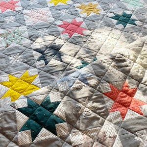 May include: A grey quilt with a patchwork design featuring various coloured stars. The stars are made of different fabrics with patterns like polka dots, floral prints, and solid colours. The quilt is quilted with a diamond pattern.