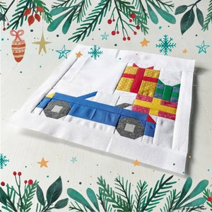 Puede incluir: Un cuadrado de tela blanco con un coche azul y regalos coloridos sobre él. El coche lleva los regalos. El cuadrado de tela está decorado con un patrón floral verde y rojo.