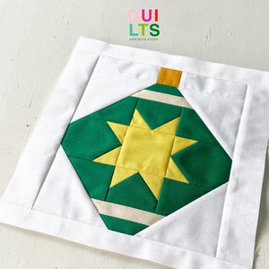 May include: A quilted Christmas ornament block featuring a green and yellow star design. The ornament has a gold top and is set against a white background with a white border. The text "QUILTS AND NICE STUFF" is in the upper left corner.