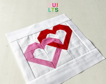 May include: A white fabric square with two overlapping hearts made of pink and red fabric. The hearts are sewn together to create a quilted design. The fabric square has a white border.