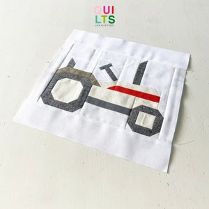 May include: A fabric quilt block featuring a tractor design. The tractor is constructed from various fabric pieces in shades of gray, white, and red. The block is set against a white background, with the text "QUILTS AND NICE STUFF" in the upper left corner.