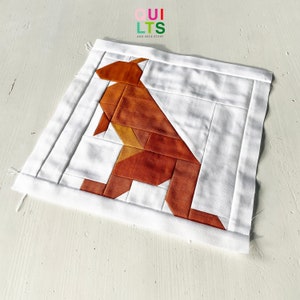 May include: A white fabric square with a brown and orange geometric kangaroo design. The fabric is quilted and has a white border.