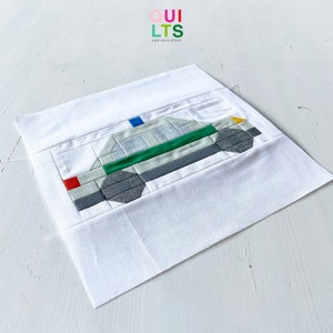 May include: A white fabric square with a green, grey, white, blue, red, and yellow patchwork design of a police car. The fabric square is on a white surface.