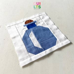 May include: A blue and white quilt block with a brown accent. The block is made of fabric squares sewn together to create a geometric design. The quilt block is a simple design with a modern feel.