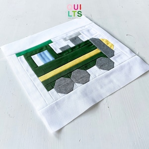 Peut inclure: Un bloc de courtepointe patchwork vert, jaune, blanc et gris représentant un train. Le bloc est sur un fond blanc.