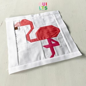 May include: A pink flamingo quilt block with a white background. The block is made up of several different pieces of fabric, sewn together to create the shape of a flamingo. The quilt block is perfect for a baby quilt, a wall hanging, or a table runner.