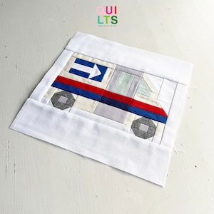 May include: A white fabric square with a patchwork design of a truck in shades of blue, red, gray, and white. The truck has a white arrow pointing to the right on the side. The fabric square is surrounded by a white border.