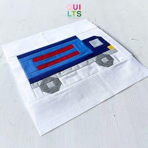 May include: A blue and red truck quilt block with grey wheels and a yellow detail. The block is on a white background.