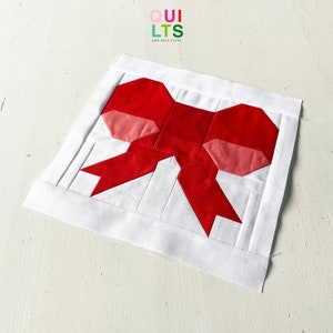 Può includere: Un blocco di trapunta a forma di fiocco rosso e rosa su sfondo bianco. Il blocco è composto da quadrati e triangoli. Il blocco di trapunta è su una superficie bianca.